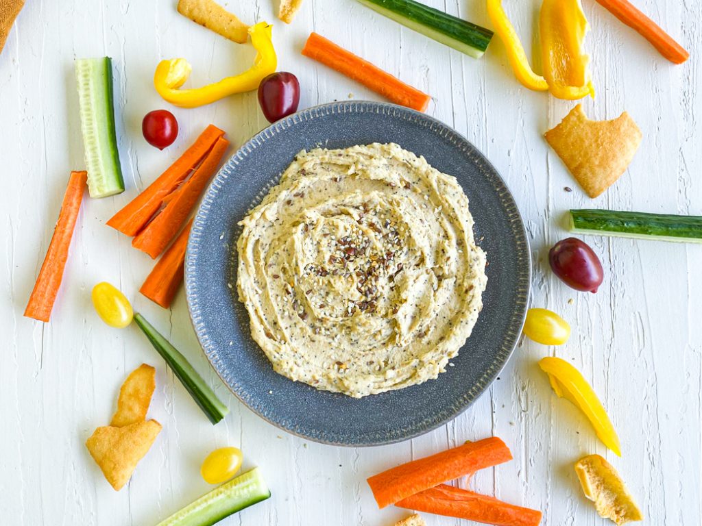 Everything bagel hummus on a plate in the center. Surrounded by raw veggies and pita chips