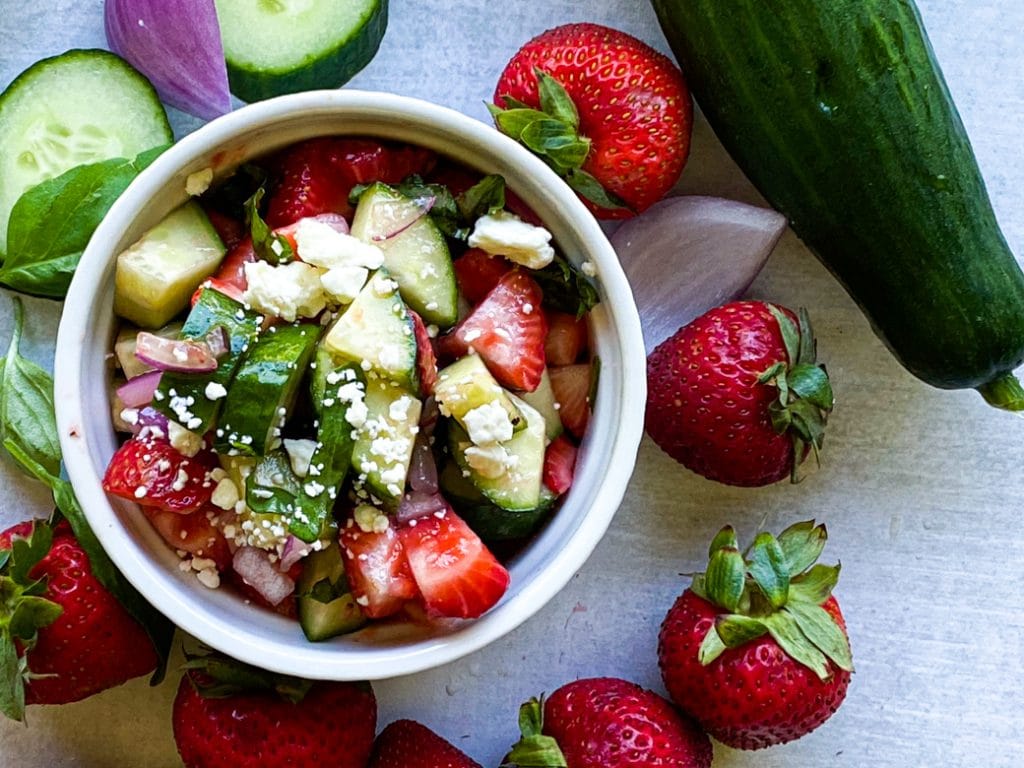 Top down view or cucumber strawberry salad in a white ramekin bowl. Sprinkled with feta cheese. Cucumber strawberry and onion on the table around the bowl. 