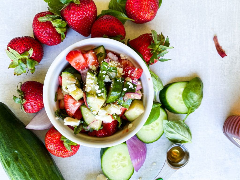 Cucumber strawberry salad in a white ramekin. Top down view. Strawberries, sliced cucumbers and onions on the table around it. 