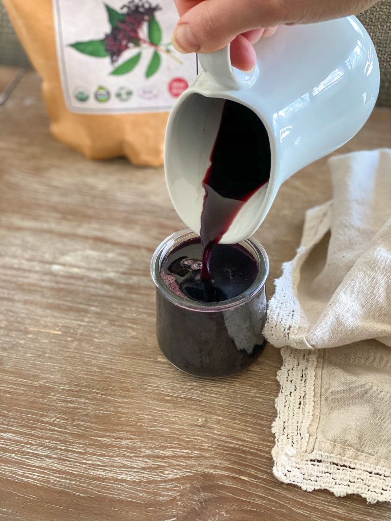 pouring elderberry  syrup into a clear glass jar. 